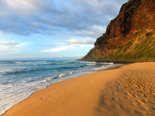 A serene beach scene with gentle waves, golden sand, and a rocky cliff under a partly cloudy sky.