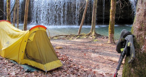 A yellow tent set up near a waterfall, surrounded by trees and fallen leaves, with a hiking pole leaning against a tree.