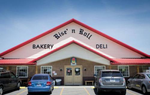 Exterior of Rise 'n Roll Bakery Deli, featuring a red roof and parked cars in front on a sunny day.