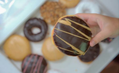 A hand holding a chocolate frosted donut with drizzled icing, surrounded by various other donuts in a box.