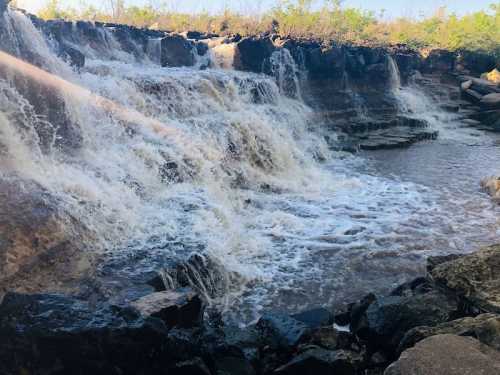 A cascading waterfall flows over rocky terrain, surrounded by greenery under a clear blue sky.