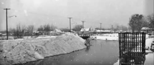 A snowy landscape with a large pile of snow by a frozen body of water, surrounded by trees and power lines.