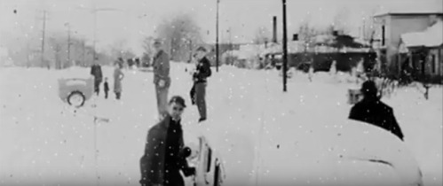 Black and white image of people in winter clothing standing in a snowy street, with snowflakes visible in the air.