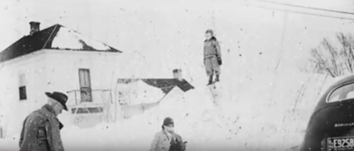 Two figures in winter clothing stand in a snowy landscape, with a house and a plane partially visible in the background.
