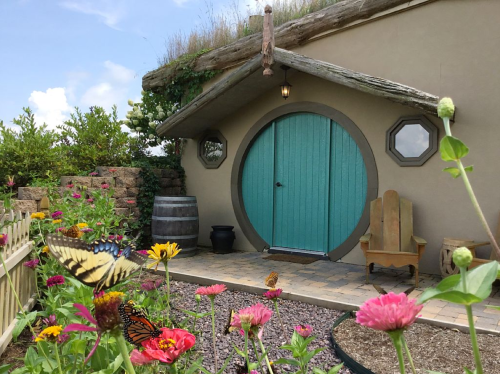 A whimsical round door with a turquoise finish, surrounded by colorful flowers and butterflies, set against a sunny sky.