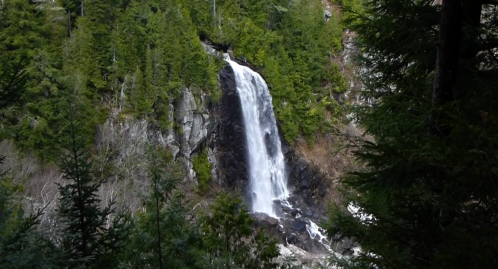A cascading waterfall surrounded by lush green trees and rocky terrain in a serene natural setting.