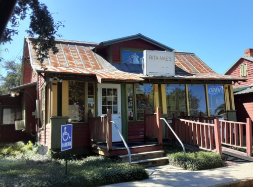 A charming red house with a metal roof, featuring a sign that reads "Rita Mae's" and an open door. Accessible parking nearby.