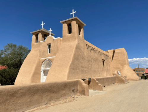 Adobe-style church with two towers and crosses, set against a clear blue sky and dirt road.