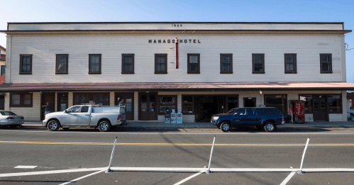 Historic building with "Manago Hotel" sign, featuring a two-story facade, parked vehicles, and a clear blue sky.