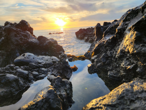 Sunset over the ocean, viewed through rocky shoreline with a small tide pool reflecting the sky.