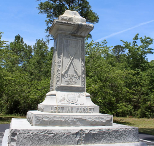 A stone monument with inscriptions, surrounded by greenery, commemorating the defenders of a historical site.