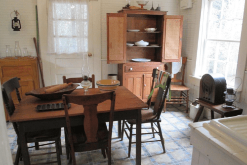 A cozy vintage kitchen with a wooden table, chairs, a hutch, and a window letting in natural light.