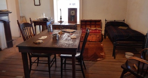 A simple, rustic room featuring a wooden table, chairs, a bed, and various vintage furnishings.