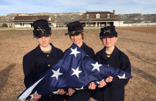 Three young individuals in military-style uniforms hold a blue flag with white stars, standing in front of a historic building.