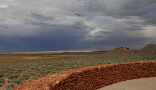 A vast desert landscape under a cloudy sky, with distant hills and a stone wall in the foreground.