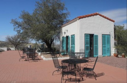 A quaint white building with turquoise shutters, surrounded by outdoor tables on a brick patio under a clear blue sky.