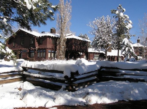 A snow-covered lodge surrounded by trees, with a wooden fence in the foreground and a clear blue sky above.