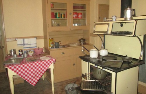 A vintage kitchen featuring a checkered tablecloth, an old stove, and wooden cabinets filled with kitchenware.
