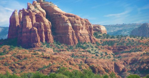 Red rock formations rise against a blue sky, surrounded by green vegetation in a scenic landscape.
