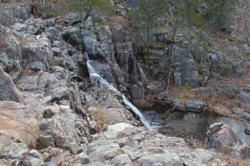 A rocky landscape with a small waterfall flowing into a pool, surrounded by sparse trees and dry foliage.