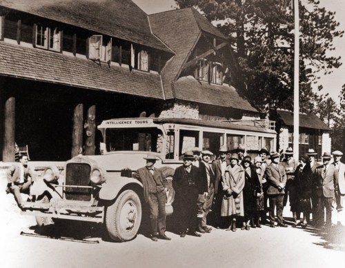 A vintage bus parked in front of a lodge, with a group of people posing for a photo in early 20th-century attire.
