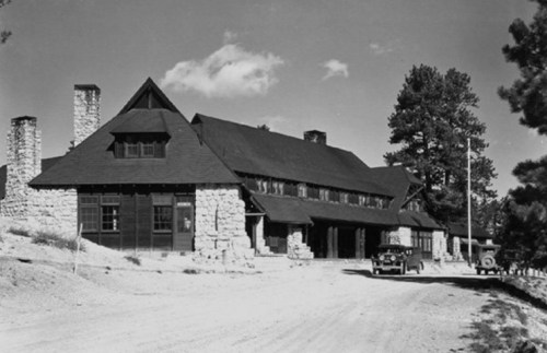 Historic stone lodge with a dark roof, surrounded by trees, on a dirt road under a clear sky. Vintage car parked nearby.