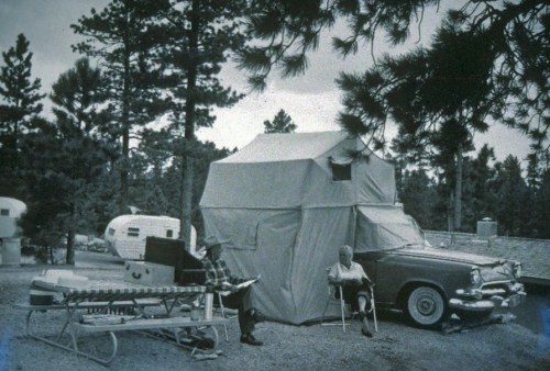 A vintage scene featuring a car and a tent, with two people sitting outside among trees and trailers.