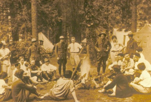 A group of people gathered around a campfire in a forest, with tents visible in the background.