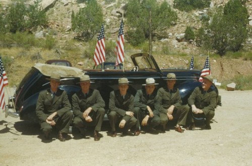 A group of six men in military uniforms poses beside a vintage convertible car adorned with American flags.