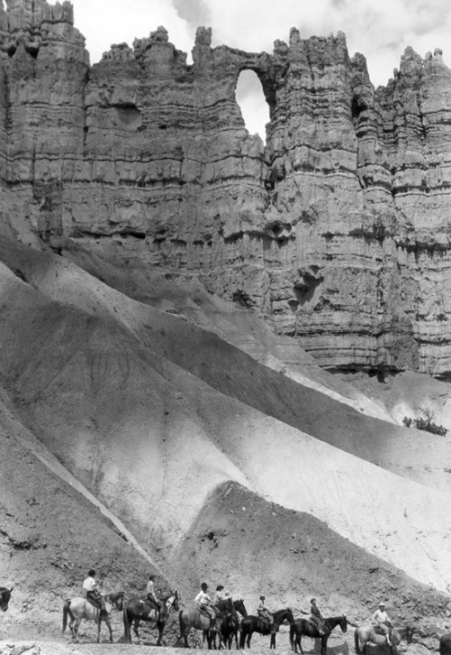 Black and white image of a group of people on horseback near a rocky formation with a natural arch.