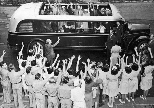 A vintage bus with passengers waving from the windows, surrounded by a crowd of people raising their arms in excitement.