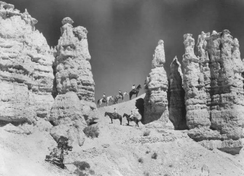 Black and white image of riders on horseback near tall, rocky formations in a desert landscape.