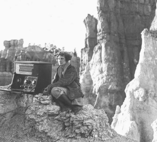A person sits on a rocky outcrop next to an old radio, surrounded by rugged cliffs in a barren landscape.