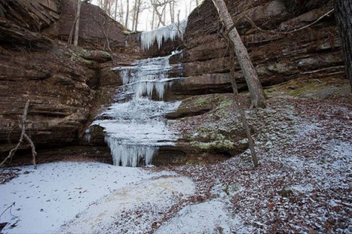 A frozen waterfall cascading down rocky cliffs, surrounded by trees and a snowy ground.