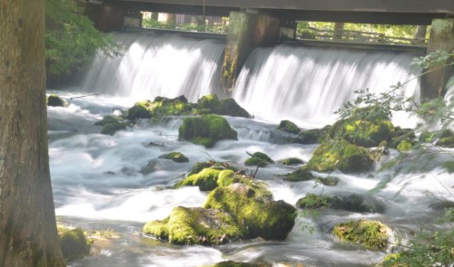 A serene river flows over moss-covered rocks under a bridge, with soft water cascading in a tranquil setting.