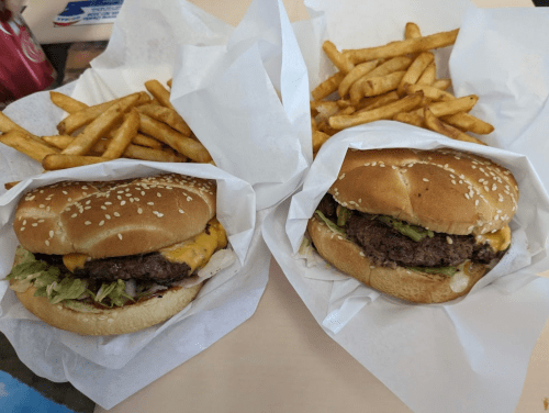 Two sesame seed burgers with cheese and lettuce, served with a side of golden French fries, all wrapped in white paper.