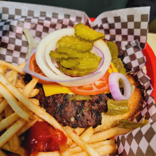 A close-up of a cheeseburger with pickles, onions, and tomatoes, served with fries and ketchup in a checkered basket.
