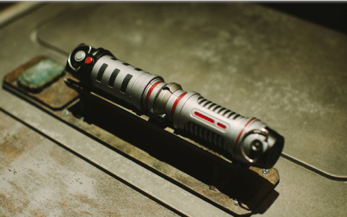 A metallic lightsaber hilt with red accents resting on a textured surface.