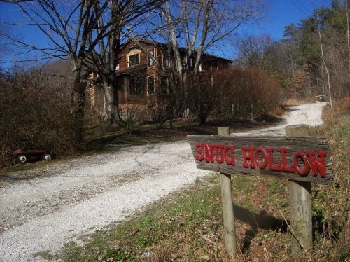 A gravel driveway leads to a rustic house, with a wooden sign reading "Smug Hollow" in the foreground.
