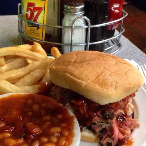 A plate with a pulled pork sandwich, baked beans, and French fries on a table with condiments in the background.