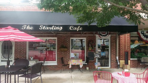 Exterior of The Starling Cafe with a black awning, red and white striped umbrellas, and outdoor seating.