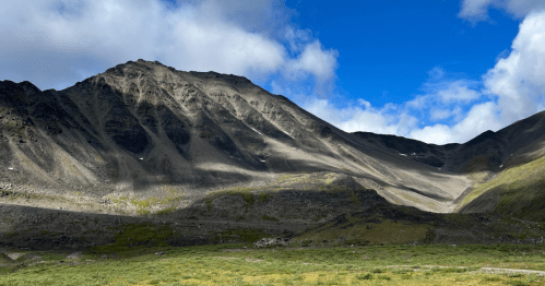 A rugged mountain landscape under a partly cloudy blue sky, with green grass in the foreground and rocky slopes.