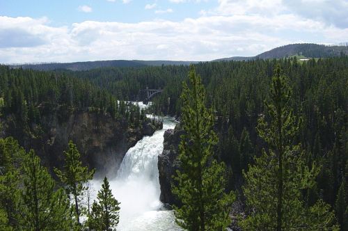 A scenic view of a waterfall cascading through a forested landscape under a partly cloudy sky.