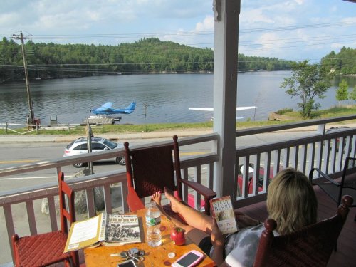 A person relaxes on a porch overlooking a lake with a seaplane and trees in the background.