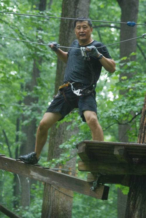 A person in athletic gear stands on a wooden platform, preparing to zip line through a lush, green forest.