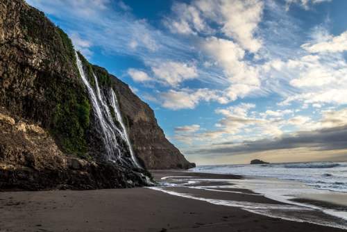 A scenic beach with a waterfall cascading down a rocky cliff under a cloudy sky. Waves gently lap at the shore.