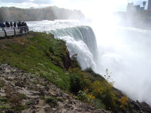 A view of Niagara Falls with mist rising, surrounded by greenery and a crowd of visitors along the railing.