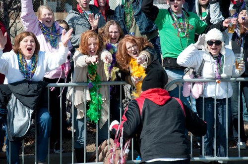 A lively crowd cheers and reaches out over a barrier, celebrating with colorful beads and festive attire.