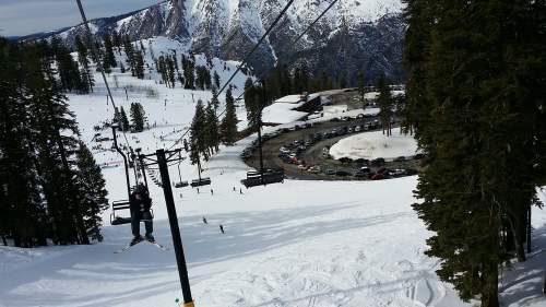 A ski lift ascends a snowy mountain, with skiers below and a parking lot visible in the background.