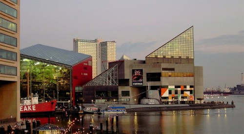 A waterfront view of modern buildings, including a glass structure and a red ship, reflecting in the water at sunset.